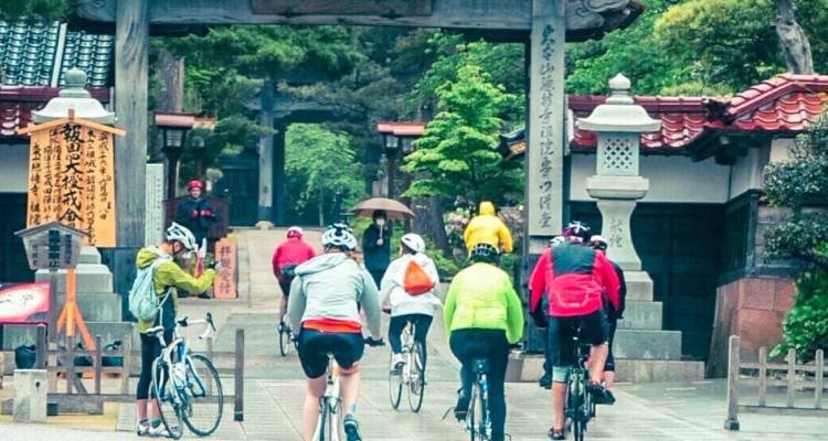 Radfahrer in farbenfroher Ausrüstung fahren durch den Eingang eines historischen japanischen Tempelkomplexes, der mit Steinlaternen und Bäumen gesäumt ist.