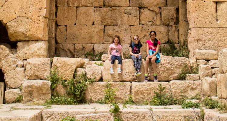 Tres niños sentados en antiguos escalones de piedra entre ruinas, sonriendo a la cámara en Jerash.
