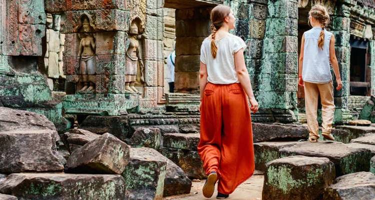 Two backpackers wander through moss-covered stone ruins of an Angkor temple complex.