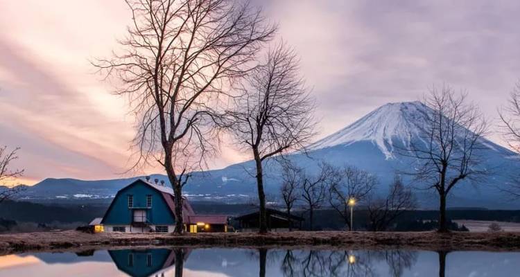 Der schneebedeckte Berg Fuji spiegelt sich bei Sonnenaufgang in stillem Wasser neben kahlen Bäumen und einem kleinen blauen Häuschen.