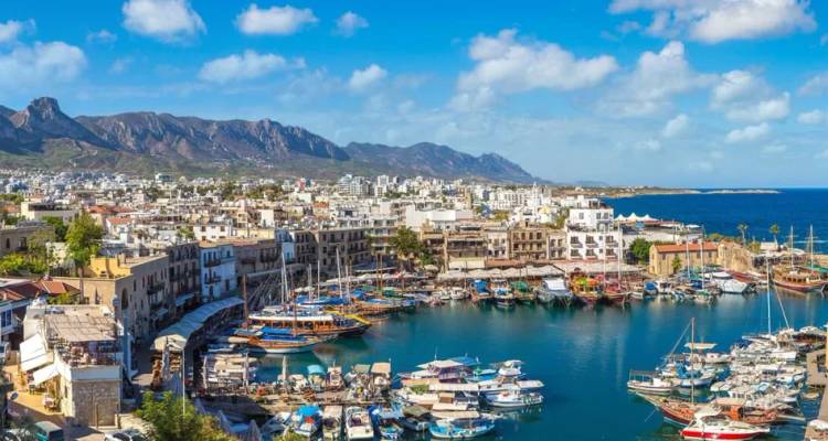 Picturesque harbor lined with boats and whitewashed buildings backed by rugged mountains under blue sky
