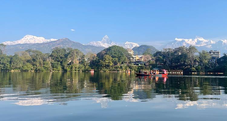 Tourist boat glides across a calm lake reflecting snow-clad Himalayan peaks and blue sky.
