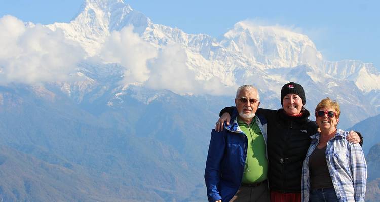 Three smiling travellers pose arm-in-arm before snow-clad Himalayan peaks under a partly cloudy sky.