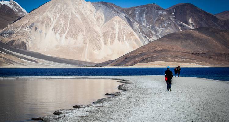 Person walking along the edge of Pangong Tso Lake with mountains.