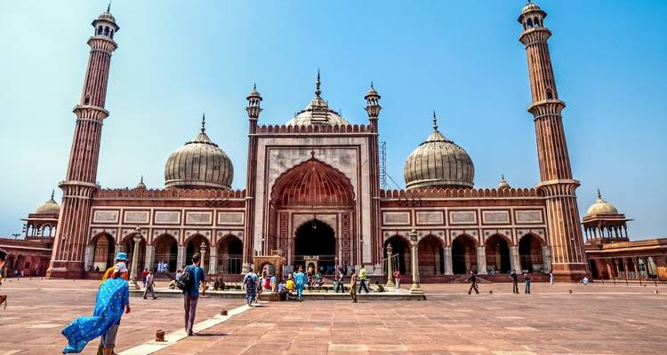 Bezoekers wandelen over de enorme binnenplaats van de Jama Masjid onder een heldere middagzon, omgeven door twee minaretten.