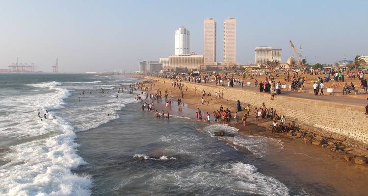 Plage bondée avec des gens, des vagues océaniques et une ligne d'horizon urbaine au loin.