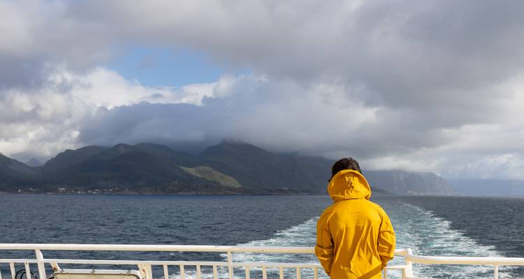 A person in a yellow raincoat on a ferry overlooking mountains.
