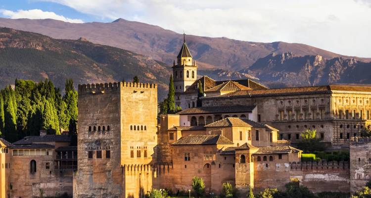 Golden-lit view of the Alhambra palace complex backed by forested foothills and distant mountains at sunset.