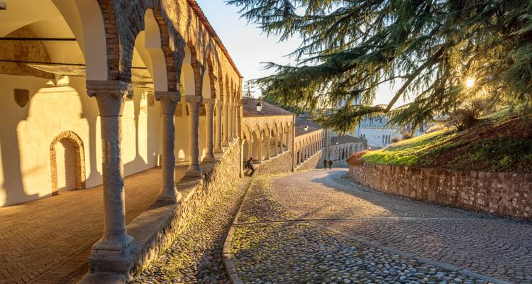 Arcada de piedra iluminada por el sol y callejón adoquinado que se curva cuesta abajo junto a un terraplén cubierto de hierba