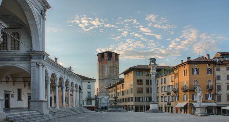 Plaza italiana histórica rodeada de un palacio con soportales, una torre medieval y fachadas en tonos pastel bajo un cielo azul