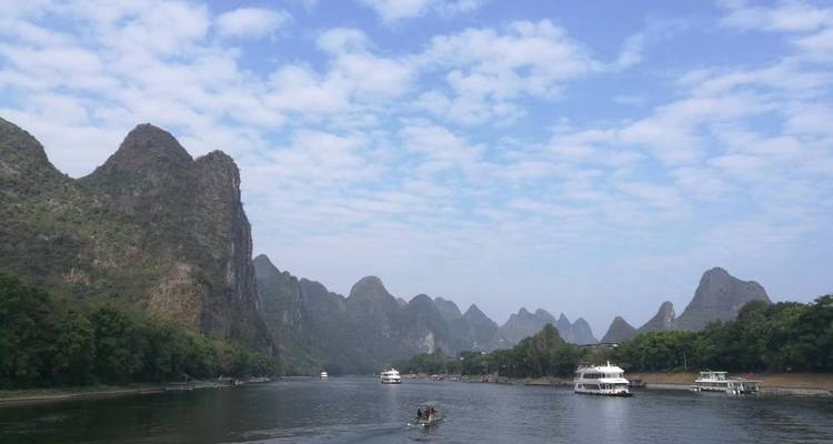 Un amplio río serpentea entre dramáticas colinas de karst bajo un cielo azul parcialmente nublado con barcos de crucero.
