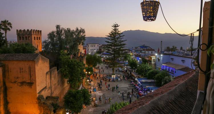Evening lights glow over Chefchaouen’s main square as the sun sets behind distant mountains.