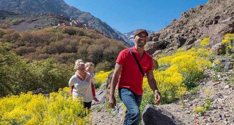 Smiling family hikes a flower-lined trail with rugged Atlas Mountains rising behind them.
