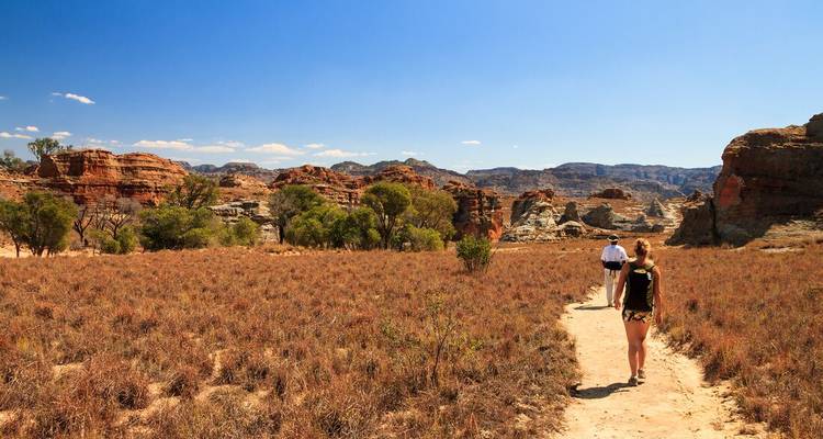Les randonneurs marchent sur un sentier poussiéreux à travers la savane aride et les affleurements de grès sous un ciel bleu dégagé.