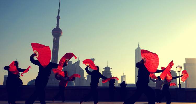 People performing a traditional dance with red fans in a cityscape.
