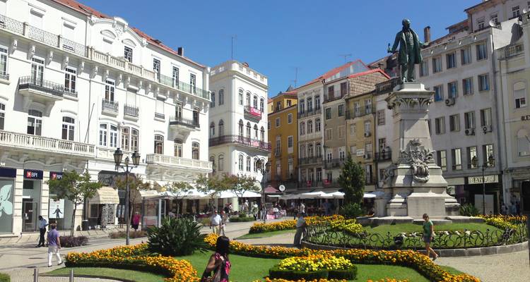 Plaza soleada del pueblo en Coimbra con parterres de flores, cafés y estatua histórica rodeada de edificios coloridos.