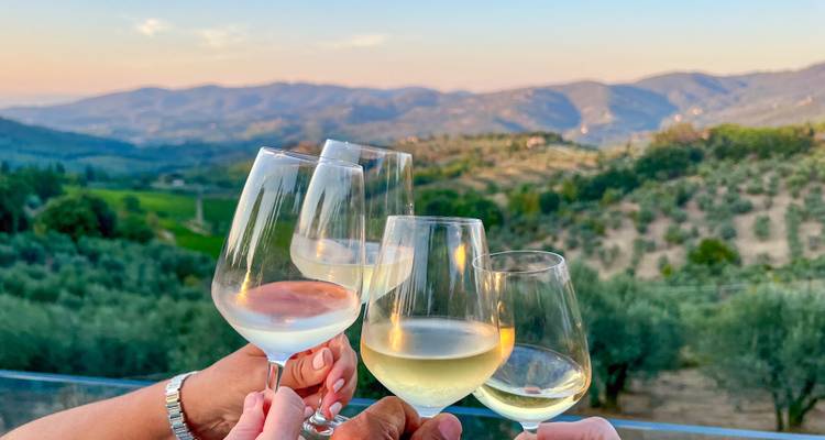 Des amis font tinter des verres de vin blanc frais devant un décor de collines toscanes ondulantes piquetées d'oliviers au coucher de soleil.