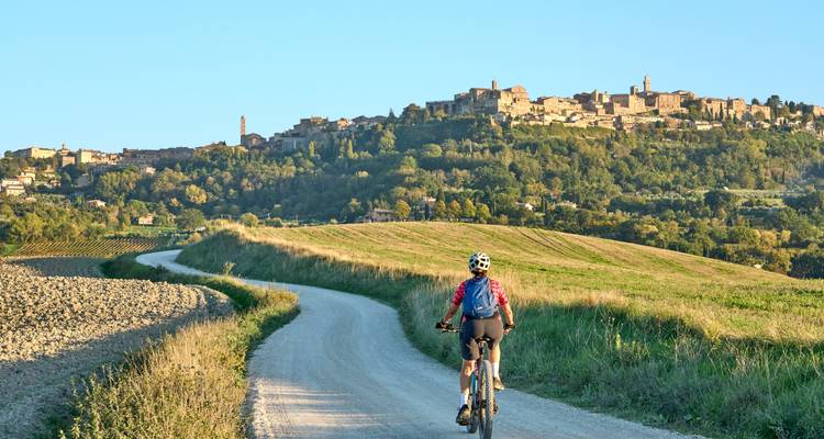 Un cycliste de montagne roule sur une route en gravier en direction d'un village toscan perché sur une colline sous un ciel bleu dégagé.
