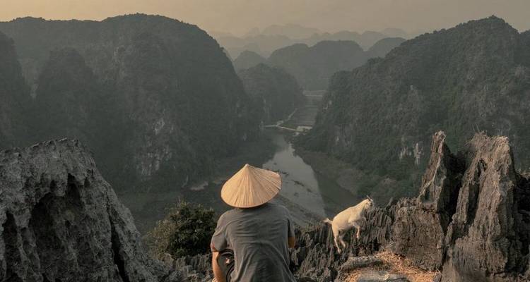Personne et chèvre sur une falaise rocheuse surplombant une vallée.