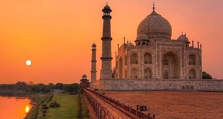 Das Taj Mahal vor einem orangefarbenen Himmel mit tief stehender Sonne am Horizont und einem ruhigen Fluss neben dem Monument.