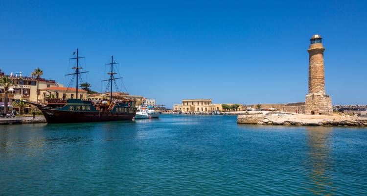 El puerto antiguo de Rethymnon con un faro de piedra bien alto, aguas azules tranquilas y una réplica de un galeón de madera.