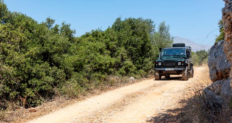Un Land Rover verde oscuro conduce por una pista polvorienta entre arbustos mediterráneos densos en un día despejado.