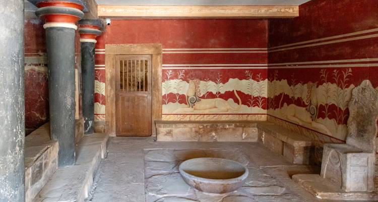 Interior de la sala del trono en el Palacio de Knossos con paredes con frescos rojos, bancos de piedra y una cuenca circular.