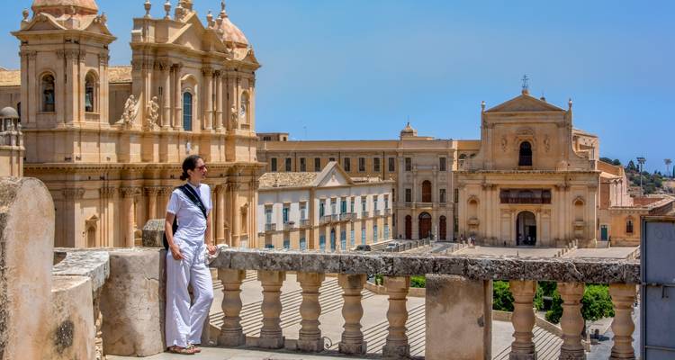 Un voyageur vêtu de blanc s'appuie sur une balustrade donnant sur la cathédrale de Noto et les bâtiments baroques environnants.