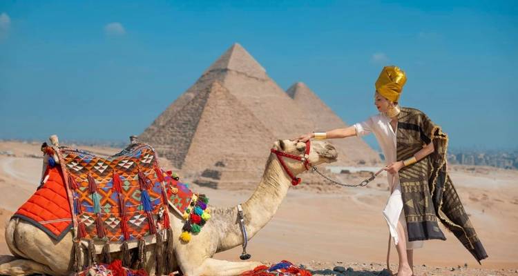 Person dressed as an ancient Egyptian next to a camel with pyramids in the background.