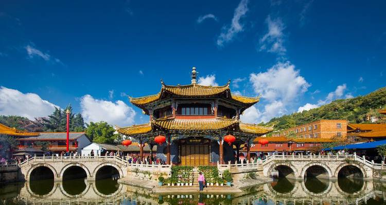 Traditional Chinese lakeside pavilion with red lanterns and arched stone bridges under blue skies.