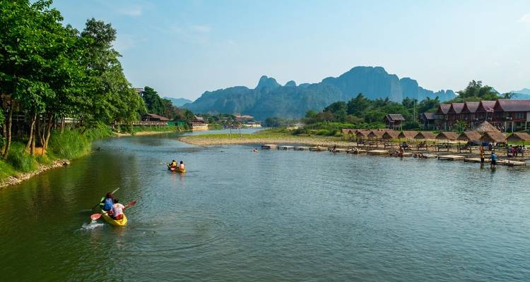 Kayakers paddle down a wide river flanked by limestone mountains near a riverside village.