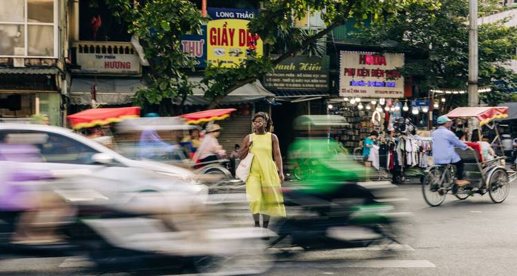 Blurred scooters zoom past a woman in a yellow dress on a busy Hanoi street.