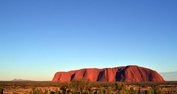 Uluru leuchtet rot im niedrigen Abendlicht über der flachen Wüstenebene.