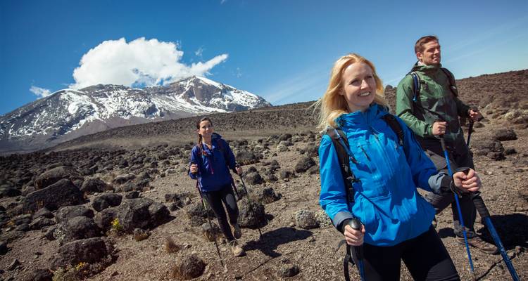 Tres trekkers con chaquetas de colores brillantes caminan por un terreno volcánico con el Kilimanjaro nevado elevándose detrás de ellos.