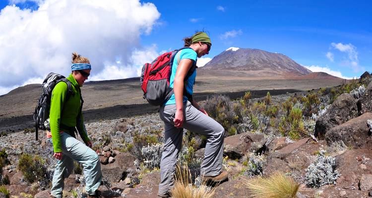 Dos excursionistas con mochilas suben por un sendero alpino rocoso con el Monte Kilimanjaro asomándose detrás.
