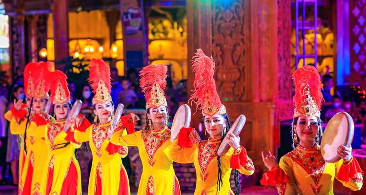 Row of dancers in bright yellow and red traditional Uyghur costumes perform energetically on a lit stage.