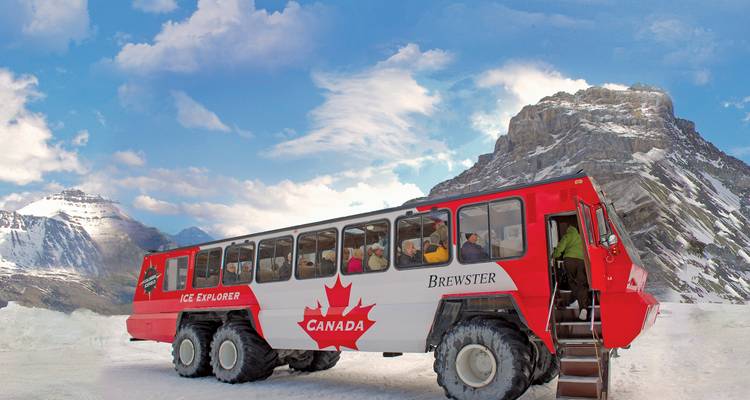A massive Ice Explorer vehicle with Canada maple leaf livery prepares to drive across a glacier under blue skies.