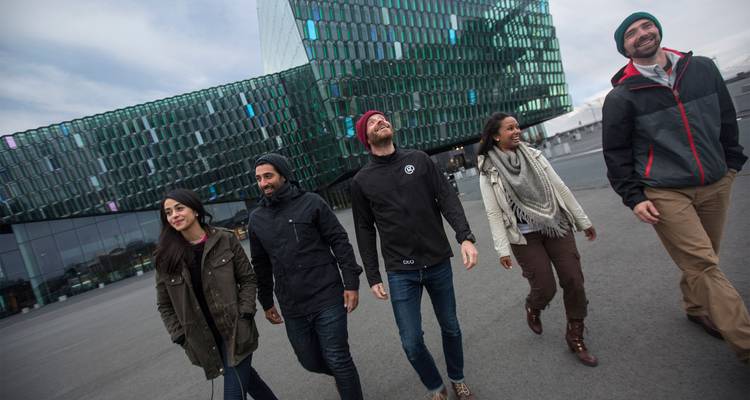 Cinco jóvenes viajeros caminan y ríen frente a una moderna sala de conciertos con paneles de cristal.