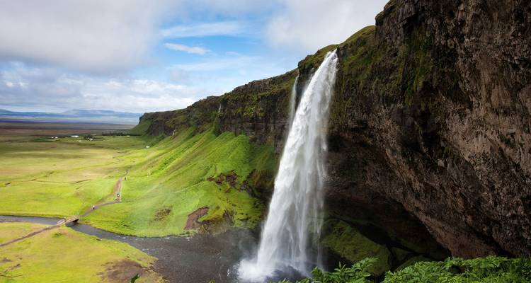 Una cascada alta y elegante que cae desde un acantilado cubierto de musgo hacia un valle verde bajo un cielo despejado.