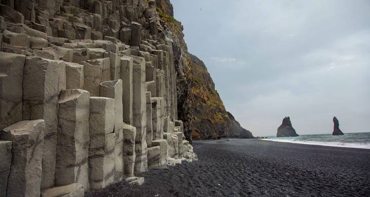 Columnas de basalto dramáticas bordeando una playa de arena negra con formaciones rocosas elevándose en la distancia.