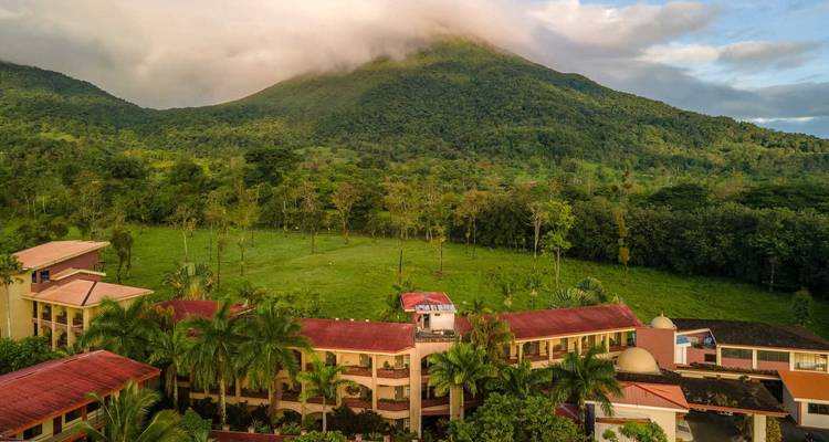 Les bâtiments de la station balnéaire se situent en dessous d'un volcan vert couronné de nuages au milieu de plaines forestières.