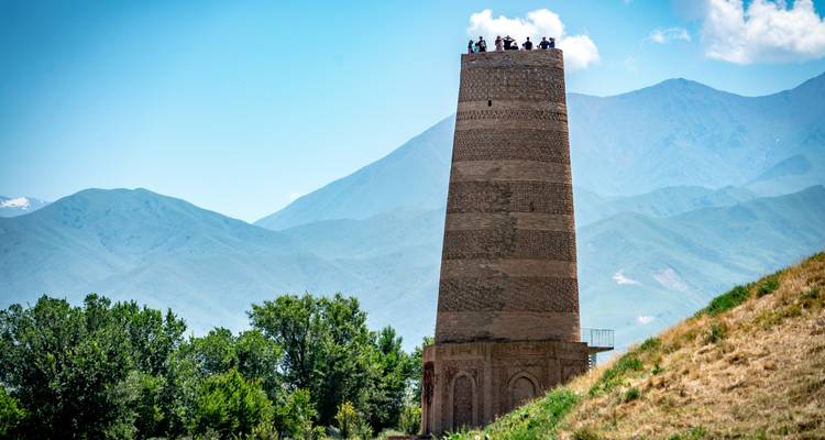 La histórica Torre Burana de ladrillo se eleva contra un fondo de árboles verdes y montañas distantes en Kirguistán.