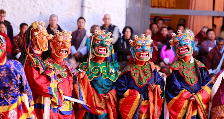 Bhutanese dancers in vivid masked costumes performing a traditional festival dance before an audience seated against a white monastery wall.