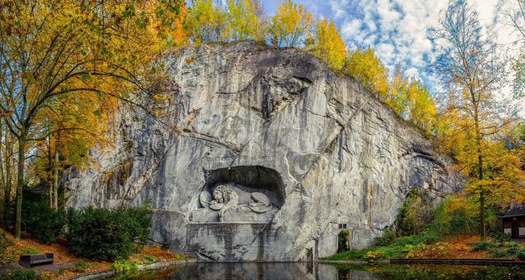 El Monumento del León de Lucerna tallado en un acantilado escarpado rodeado de follaje otoñal dorado reflejado en un estanque.