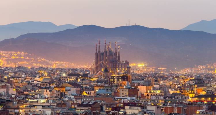 Panorama vespertino de Barcelona con la Sagrada Familia iluminada contra las montañas al anochecer.