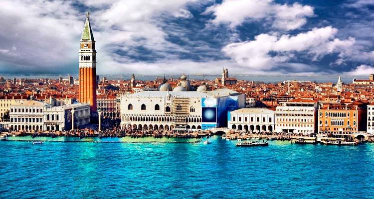 Panoramablick auf Venedig mit dem Campanile di San Marco, dem Dogenpalast und roten Ziegeldächern vor den leuchtend türkisfarbenen Lagunengewässern unter einem teilweise bewölkten Himmel.