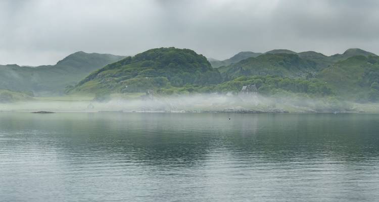 Misty highland lake surrounded by rolling green hills under grey sky