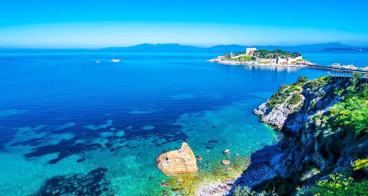 Mar Egeo turquesa vibrante con una pequeña isla fortificada y costa rocosa bordeada de flores bajo un cielo azul despejado.