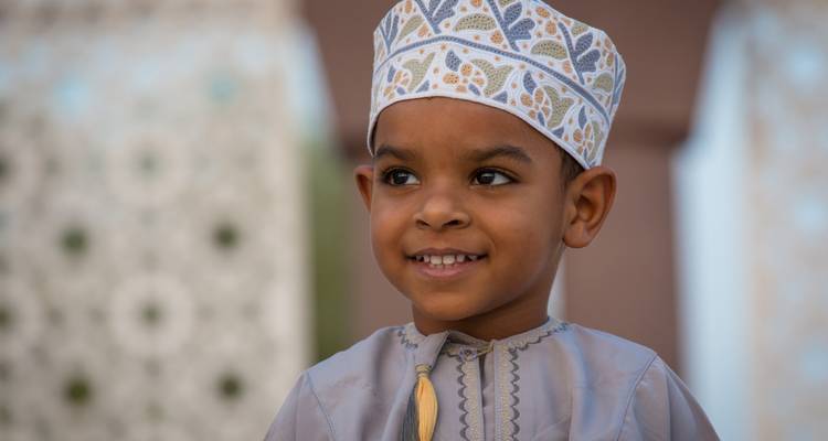 Retrato de un niño omaní sonriente usando un gorro bordado tradicional contra un fondo suave.