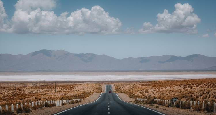 Un camino de asfalto recto que atraviesa un amplio desierto de salares hacia cadenas montañosas lejanas bajo nubes dispersas.